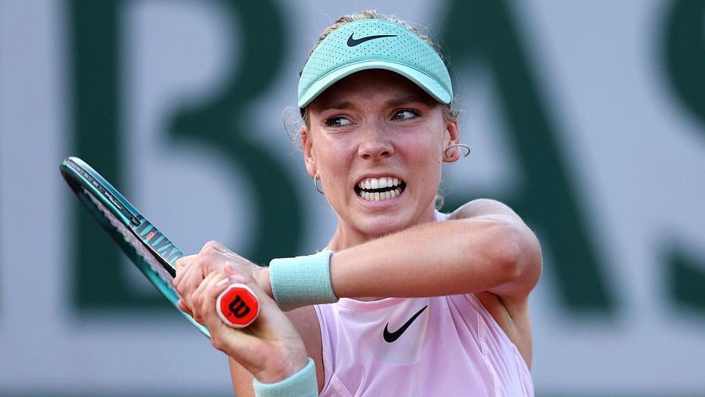 Katie Boulter, wearing a lilac sleeveless top and mint visor, grimaces as she plays a backhand, with her racquet held in two hands over her right shoulder