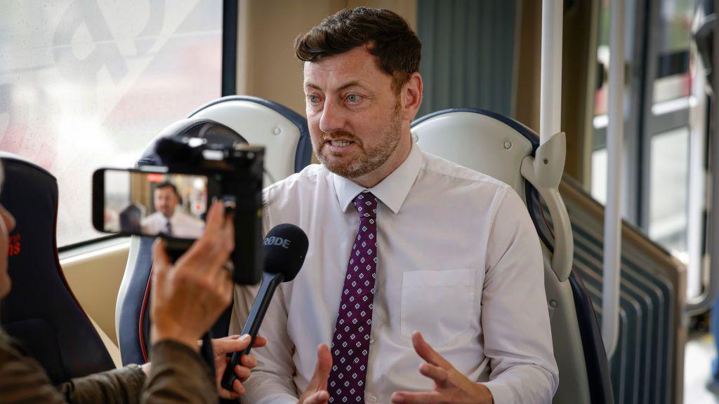 Cammy day in a shirt and tie sitting on a tram seat being interviewed by a woman holding up an iphone and a microphone