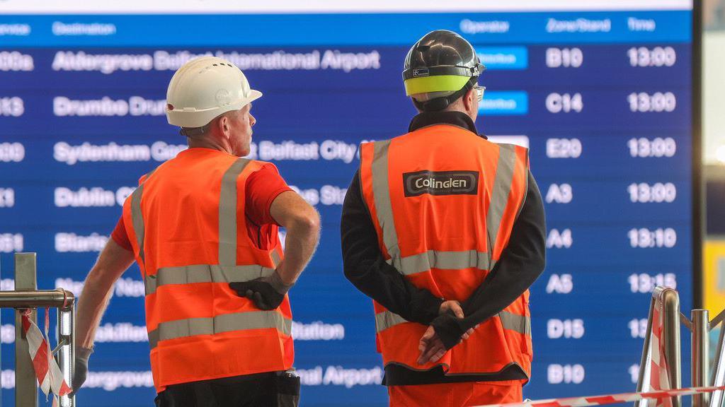 Two men wearing hi-vis vests and hard hats looking at a sign showing bus departures 