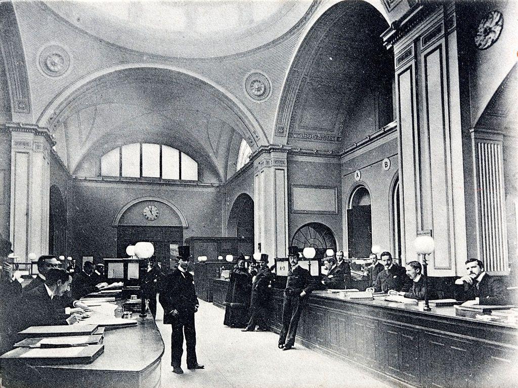 Black and white photo of one of the offices in the Bank of England in 1910 with men sitting behind wooden counters and others wearing top hats standing beside them