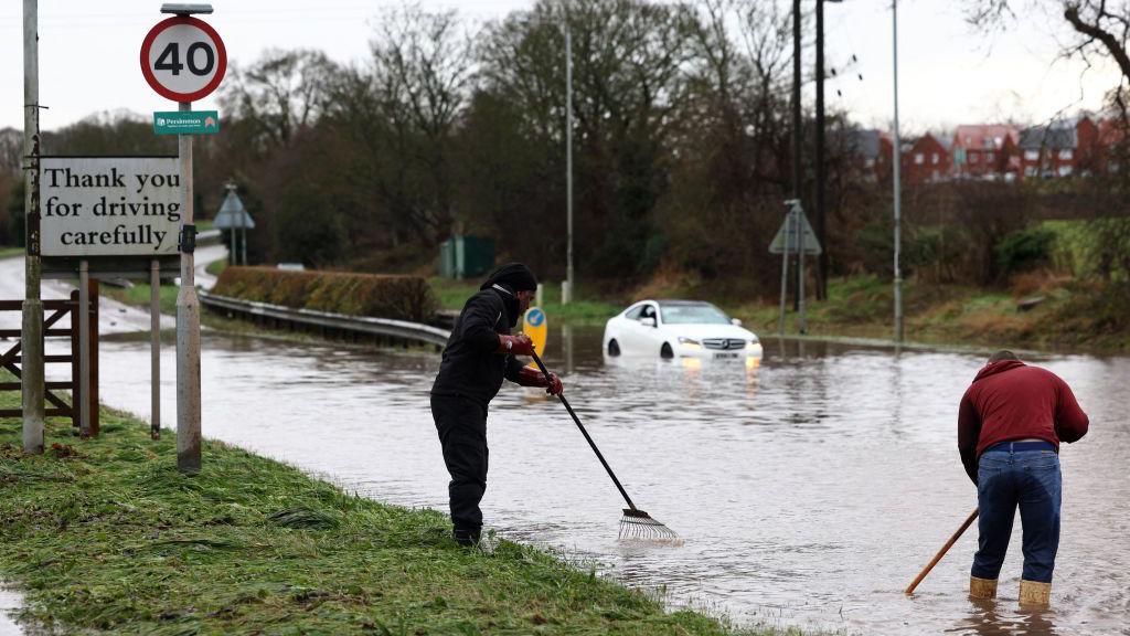 A flooded road with two people with sticks and rakes trying to unblock a drain with a white car in the background and a sign in the foreground reading 'thank you for driving carefully), in Hather, Leicestershire in January.