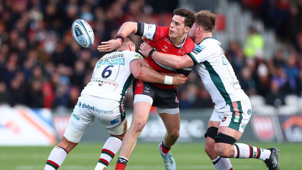 Rugby player in red offloads rugby ball as he is tackled by two opponents wearing white and green