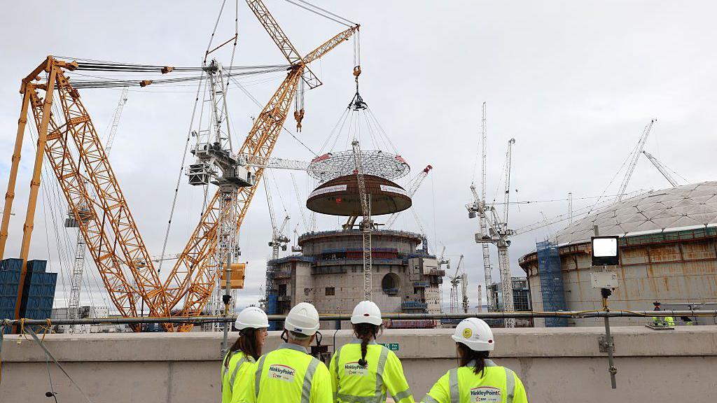 A media crew gathers to witness the world's largest land crane 'Big Carl' lifting a 245-tonne domed roof onto Hinkley Point C's reactor building in the background of the image