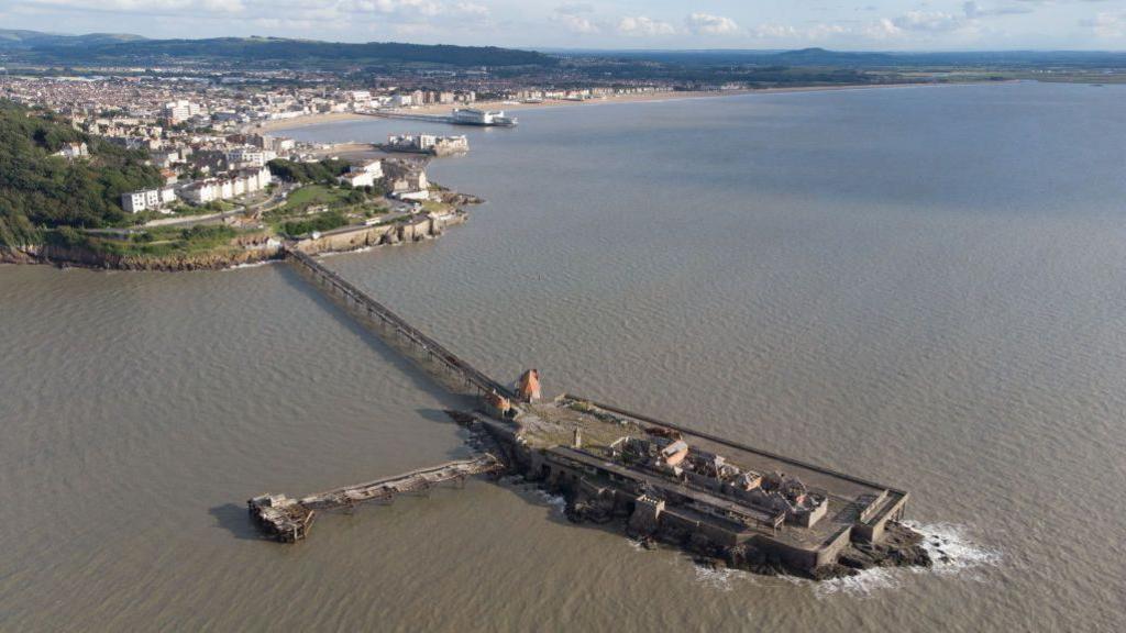 Aerial view of pier from the sea side, looking back towards the land. Pier is rectangular with some dilapidated buildings on top, and a second smaller pier coming off it to the left. The main structure is connected to the land by a narrow walkway of around 300m.