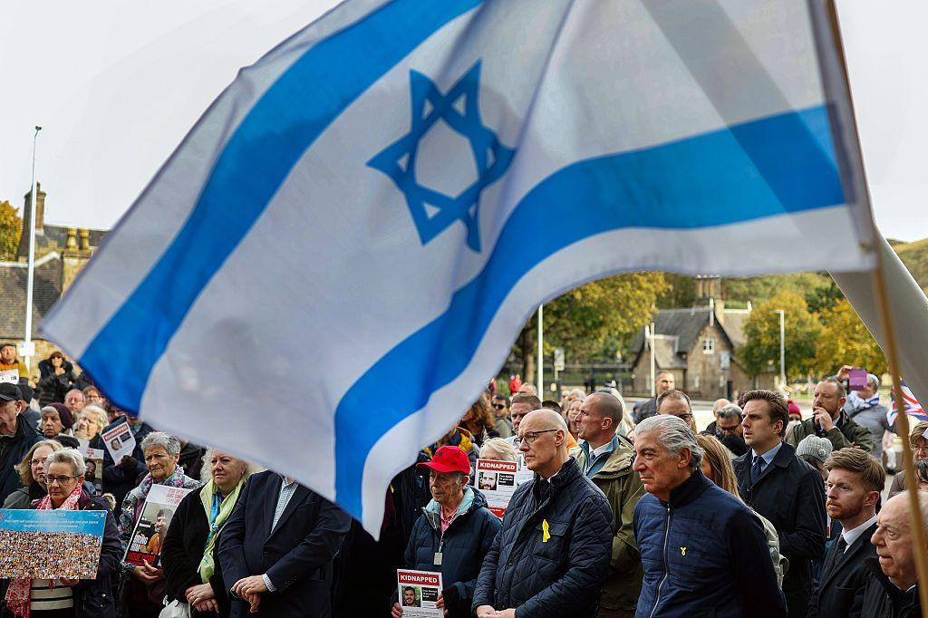 A large Israeli flag can be seen dominating much of the frame. Below, First Minister John Swinney, in a dark blue jacket, is standing among a group of people holding signs about the kidnapped victims of the 7 October attacks. They are standing outside the Scottish Parliament and Holyrood Park can be seen in the background.