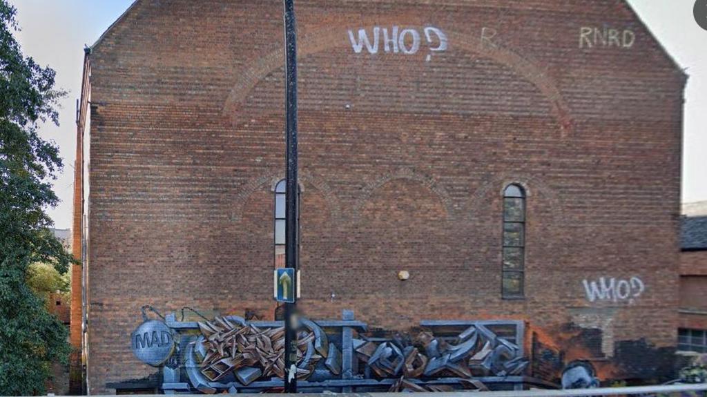 Large brick wall at rear of the Corn Exchange hall with two very narrow windows. There is graffiti at the base and the word "Who?" sprayed twice onto the wall.