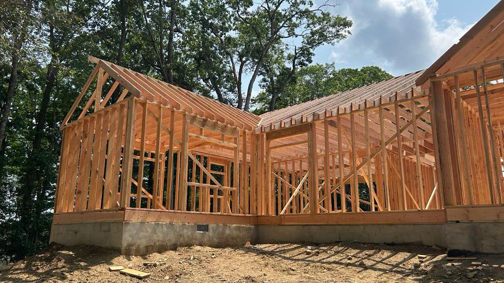 A house under construction, with only the wooden frame built, on a dirt surface. next to trees against a blue cloudy sky.