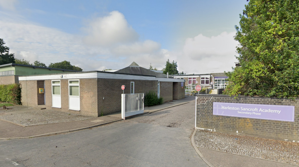 The outside of Harleston Sancroft Academy featuring a low brown-bricked building, an open white gate and a sign that reads "Harleston Sancroft Academy, Secondary Phase'. 