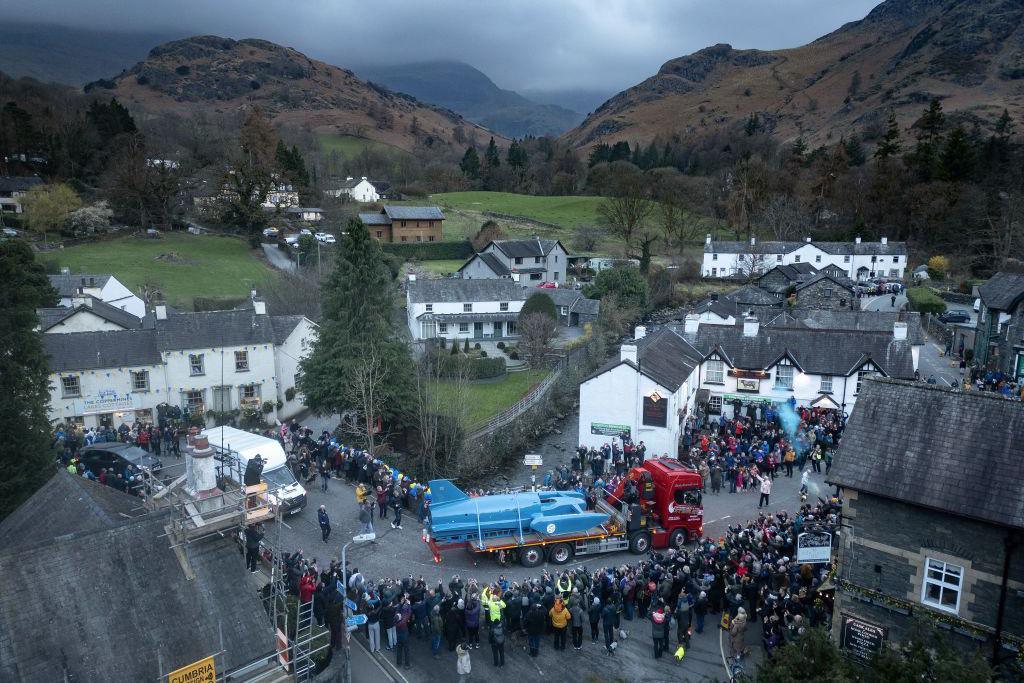 Bluebird, a jet-powered craft, arrives in Coniston secured to the trailer of a lorry. It is painted blue. Crowds line the village's narrow streets. A number of white houses and businesses stand next to the roads. Lake District fells can be seen in the background.