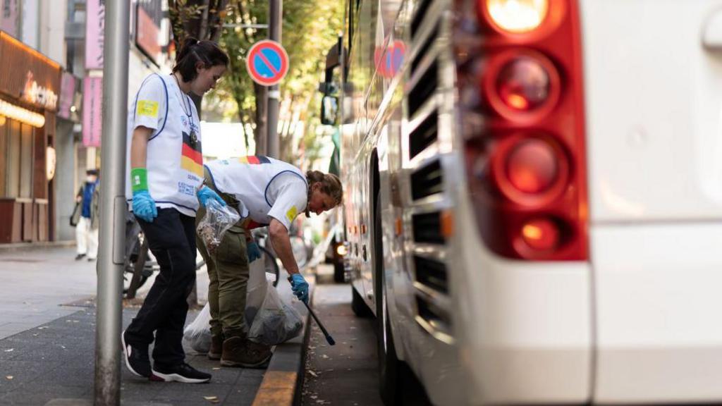 two people in white bibs with a German flag on them picking up litter by a bus on the road