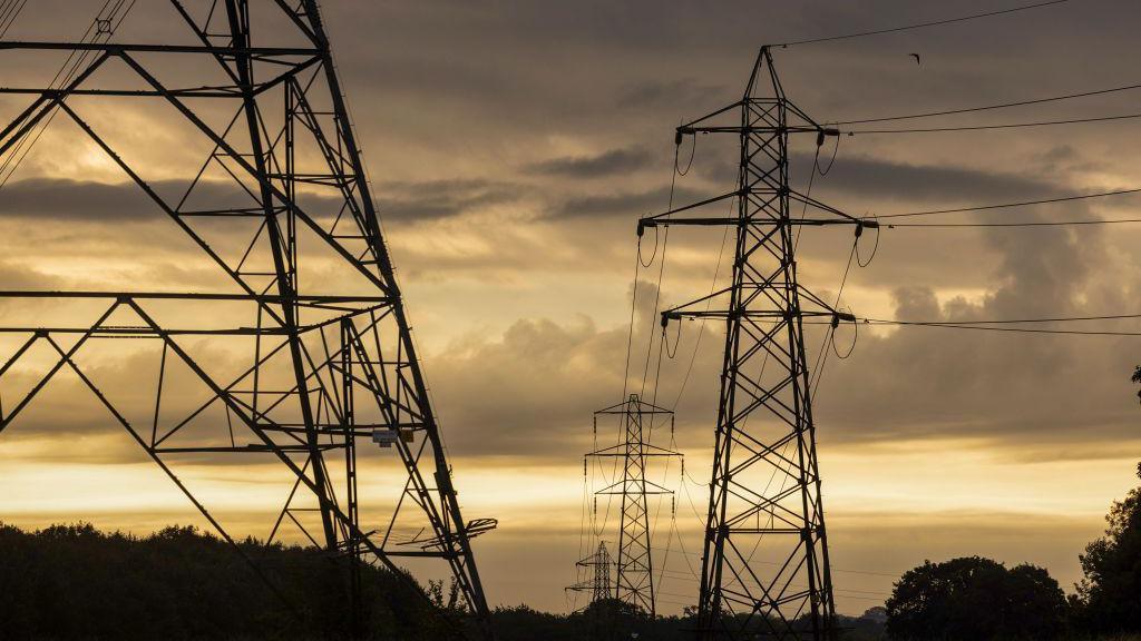 Several large metal electricity pylons are towering above trees against the sunrise. The clouds are grey with yellow light filtering through, and trees and bushes are silhouetted.