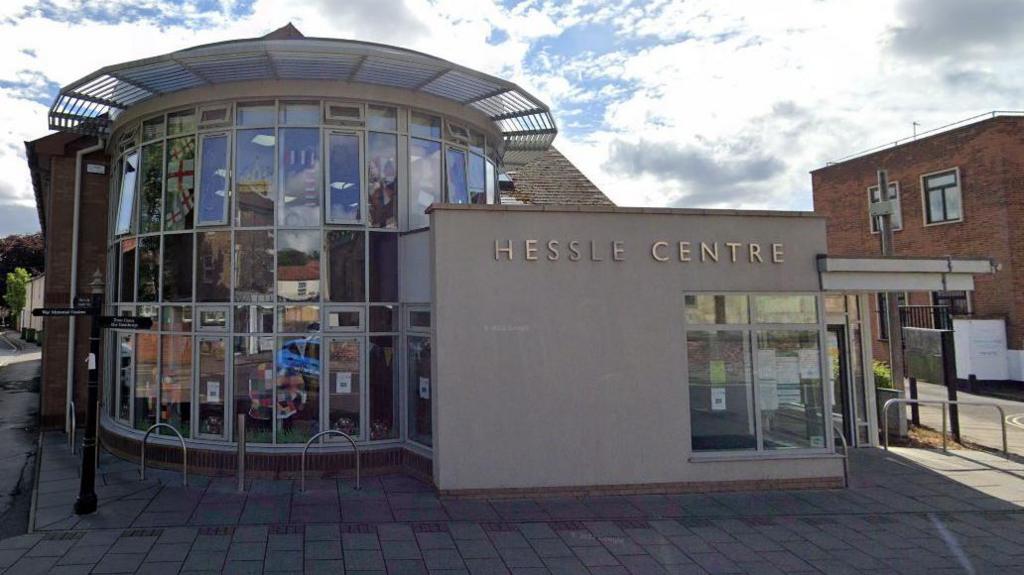 Hessle Library and Community Centre. The left half of the building has a mainly-glass circular frontage. The right-hand side is the entrance which has HESSLE CENTRE in gold lettering towards the top of the wall. 