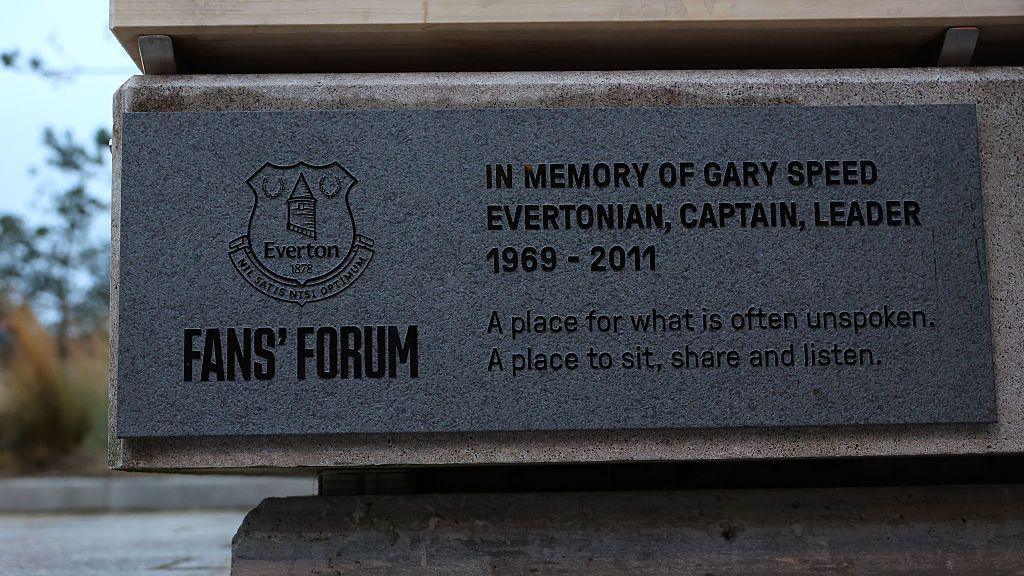 View of plaque on the side of the bench. It says "In memory of Gary Speed - Evertonian, captain, leader - 1969-2011. A place for what is often unspoken. A place to sit, share and listen". 