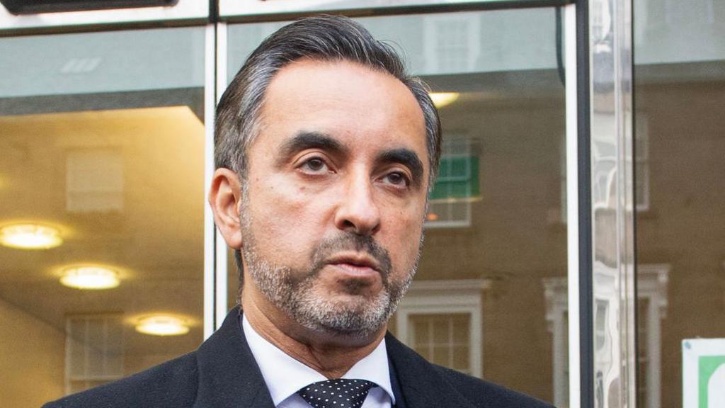 Headshot of solicitor Aamer Anwar. He has a beard, is wearing a suit and a dotty tie. He is standing in front of glass panelled doors.  