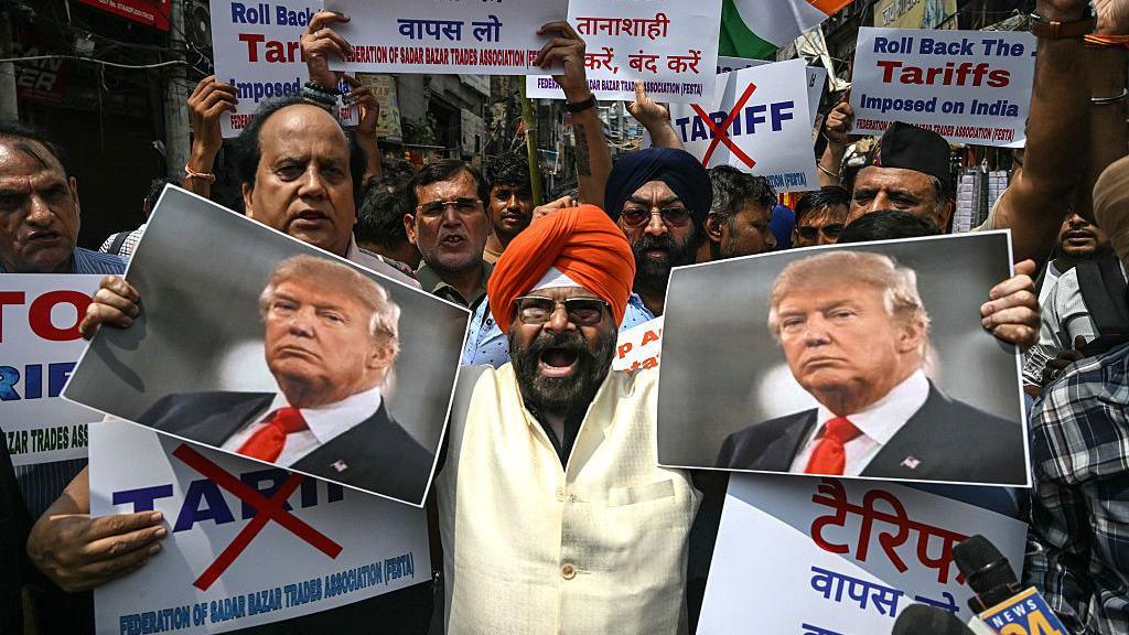 A man wearing an orange turban and white top holds up two pictures of Donald Trump and shouts angrily at the camera. Behind him are a crowd of men holding up signs stating "roll back the tariffs imposed on India".