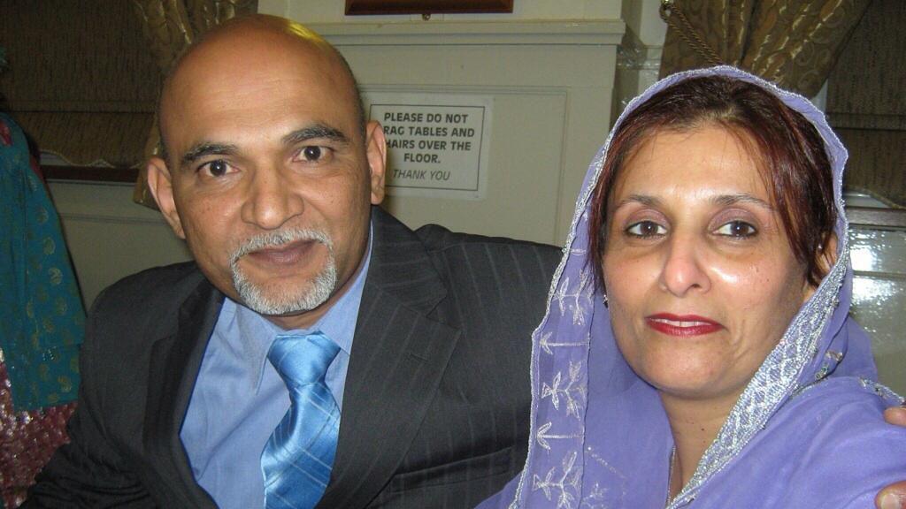 A bald man in a suit puts his arm around a woman in purple cloth loosely covering her hair and shoulders.