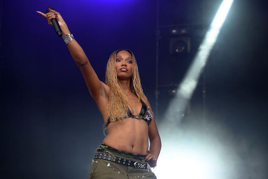 A woman wearing combat trousers and a bikini-style top waves her microphone in the air.