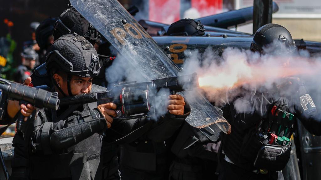 A police officer clad in black protective body armour fires a tear gas canister while other officers stand next to him with shields