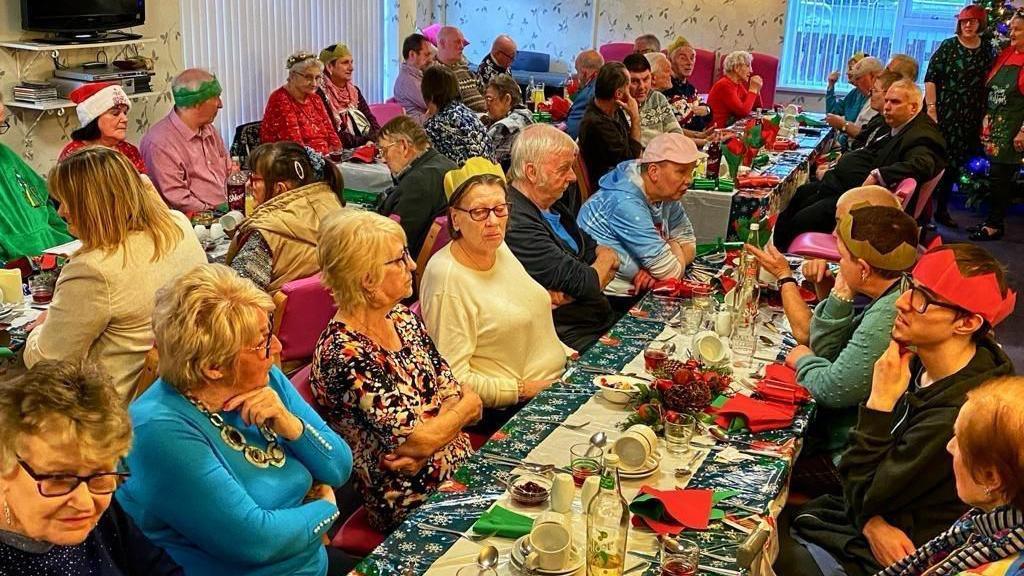 A previous Christmas Day dinner organised by Margaret Peacock in Coleraine.  Dinner guests from a mix of ages are sitting and chatting around long tables, some are wearing paper party hats, tinsel or Santa hats. 