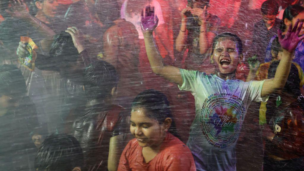 A boy closes his eyes and jumps for joy wearing a happy holi t shirt as he's sprayed with water, his hands are covered in purple paint