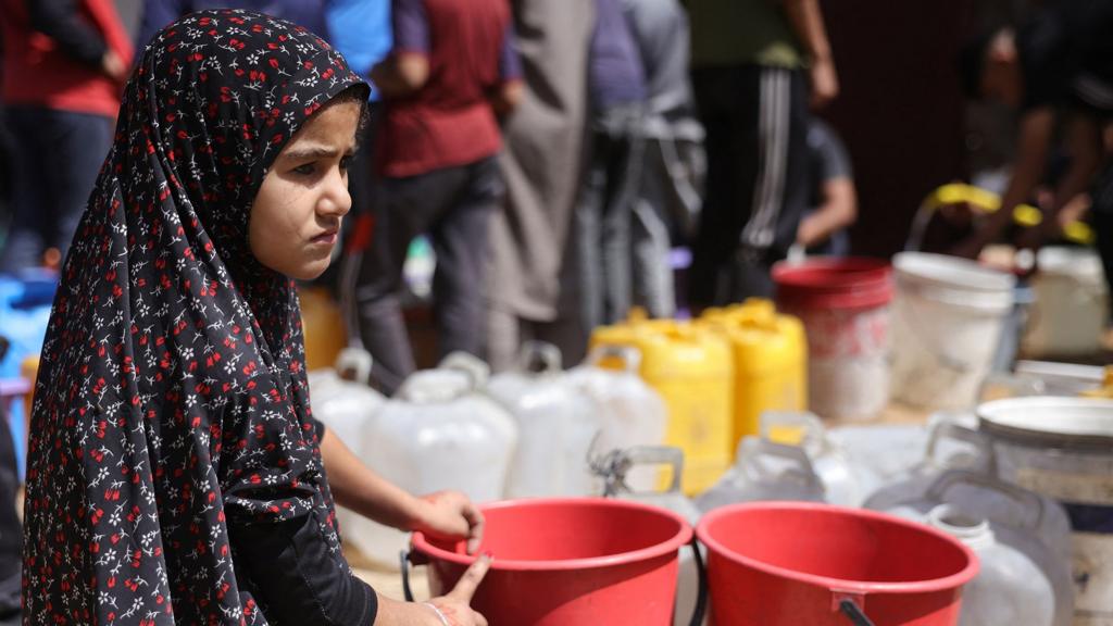 A young girl looks on as she holds a red bucket with gallon bottles of water next to them and people standing in a queue in the background, in Gaza City on 20 May