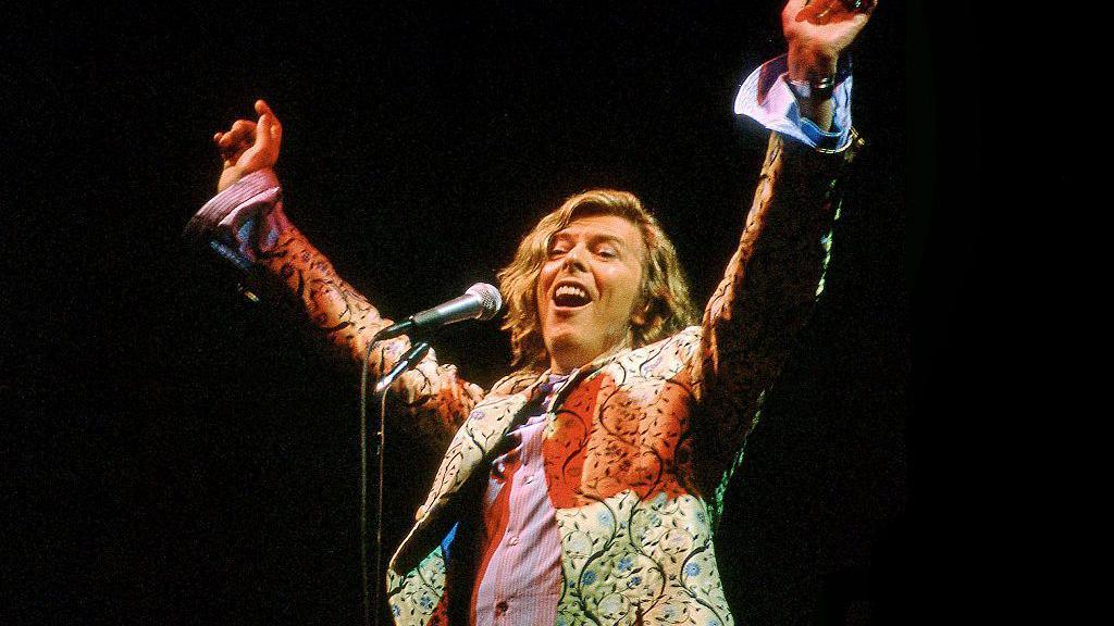 David Bowie singing into a camera with his arms outstretched above him. He is wearing an embroidered suit jacket.