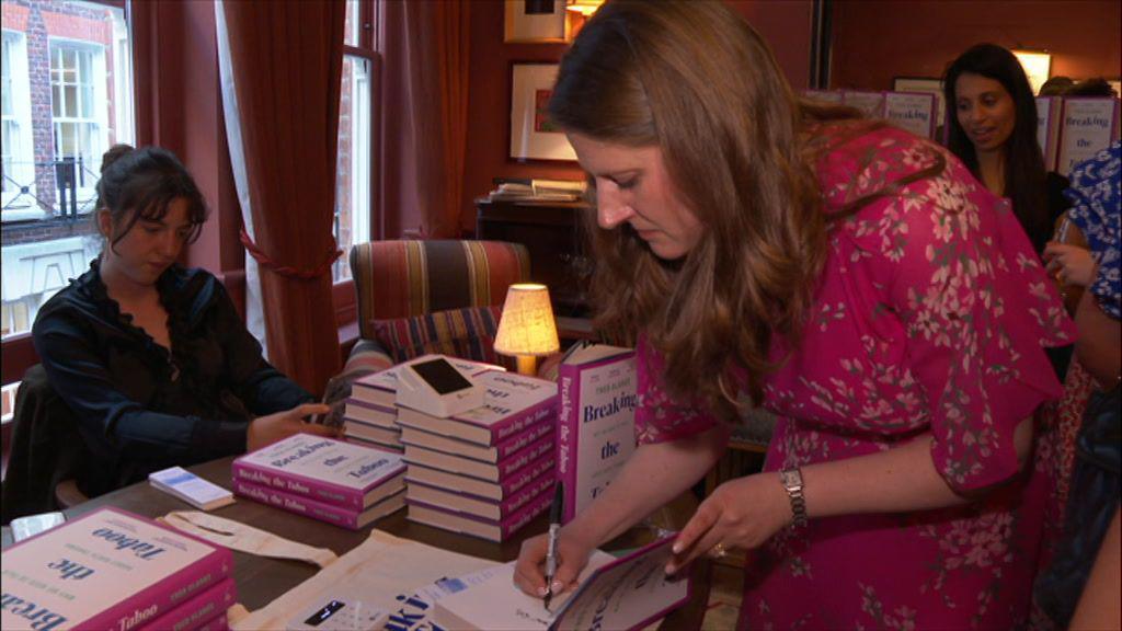 A woman with long dark hair leans over a desk to sign a book. She holds a pen in her right hand and wears a magenta dress with a flowery pattern on it. On her left wrist is a wristwatch. She signs the book which is among several piles of other books.