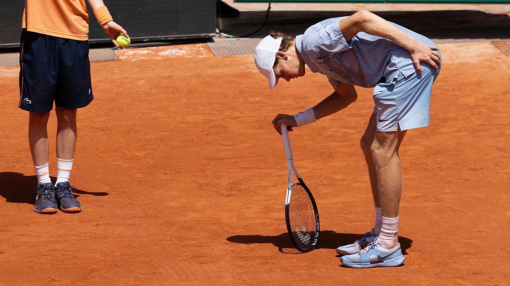 Sinner (right), wearing a grey t-shirt, shorts, and a white cap, bends over, leaning on his racquet with his right hand and clutching the top of his thigh/bottom with his left