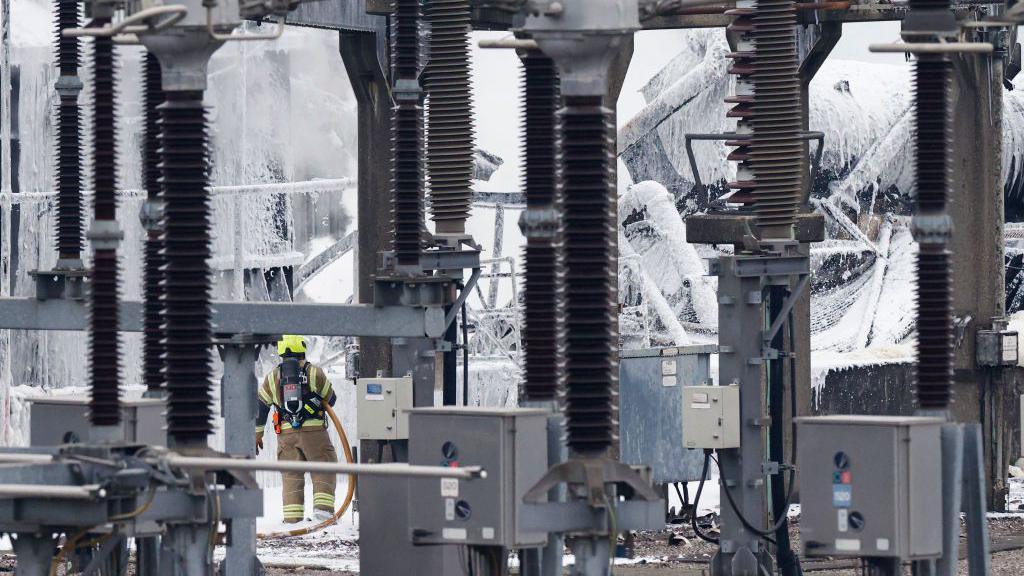 A uniformed firefighter sprays foam onto parts of the substation on Friday morning. White foam covers many parts of the substation in the background