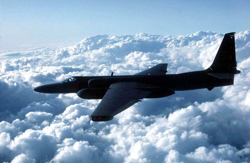 A black U-2 spy plane flying from right to left above thick clouds. 