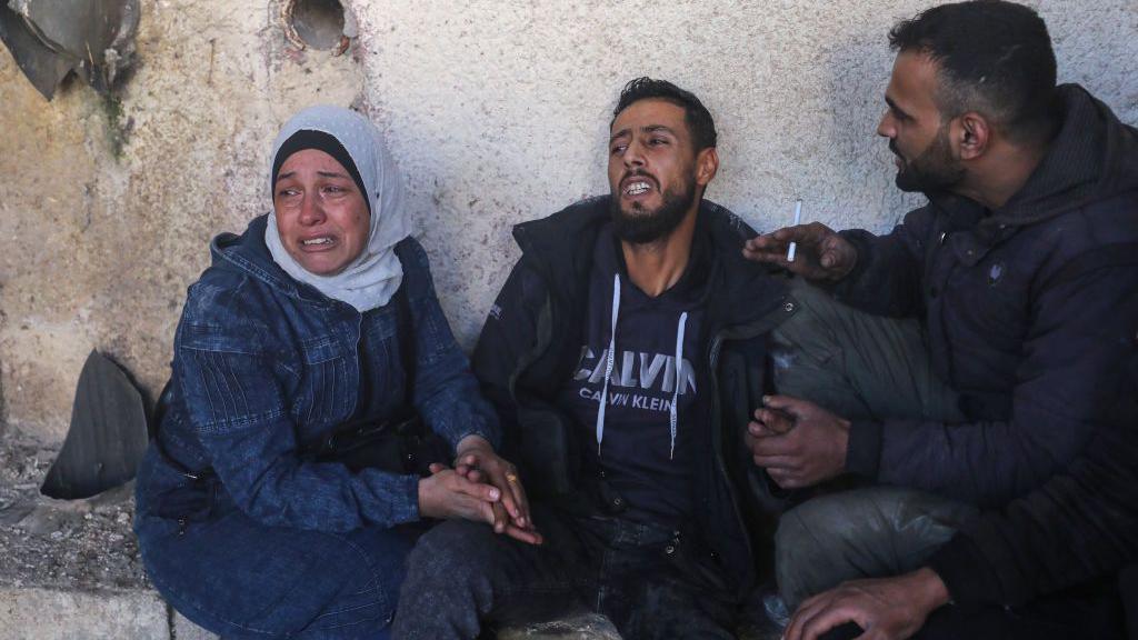 Three people sit on the ground, leaning against a wall, at a prison. A woman and man appear to be crying as they hold hands, while a man holds their hand and has his hand out as if to calm them