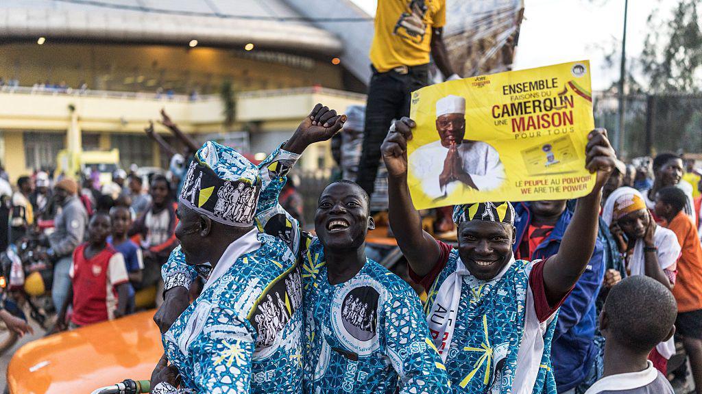 Three men, smiling and gesturing, dressed in bright and pattered matching blue outfits, hold up a sign in support of the opposition leader. There are many people behind them, in a rally.