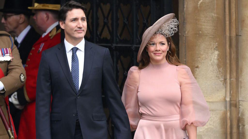 Justin Trudeau and Sophie Grégoire Trudeau arrive at Westminster Abbey for the Coronation of King Charles III and Queen Camilla on May 06, 2023 in London, England. Trudeau is wearing a navy blue suit with a polka-dotted navy blue tie, while Sophie is wearing a blush pink dress with a high neck and chiffon, billowing sleeves. She is wearing a matching fascinator on her head. The two are holding hands and smiling.