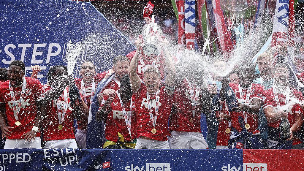 Charlton players celebrate jumping up and down with the League One play-off final trophy, shaking bottles of champagne