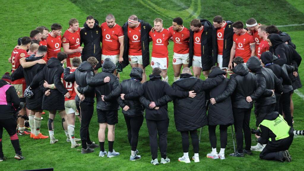 A picture of the Welsh rugby six nations squad huddling together in a circle on the pitch in the Italy vs Wales game. Some are wearing black coats and others are in their red jerseys. 