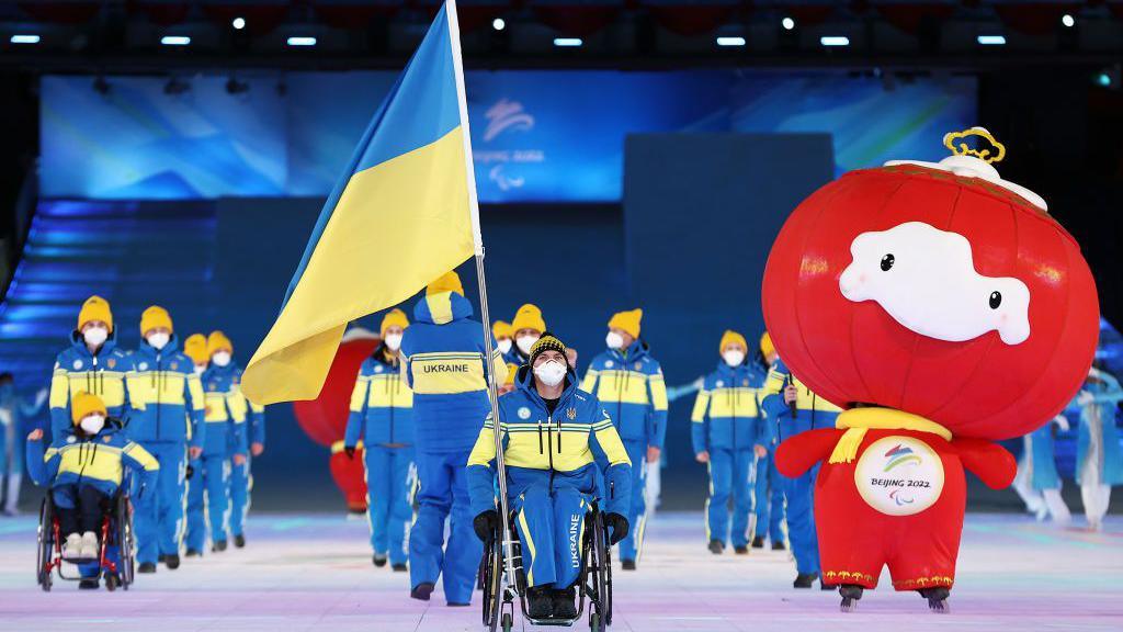 Maksym Yarovyi leads out Ukraine at the 2022 Paralympics opening ceremony in Beijing, followed by team-mates and accompanied by a Games mascot. Yarovyi is seated in a wheelchair and holds a large Ukraine flag