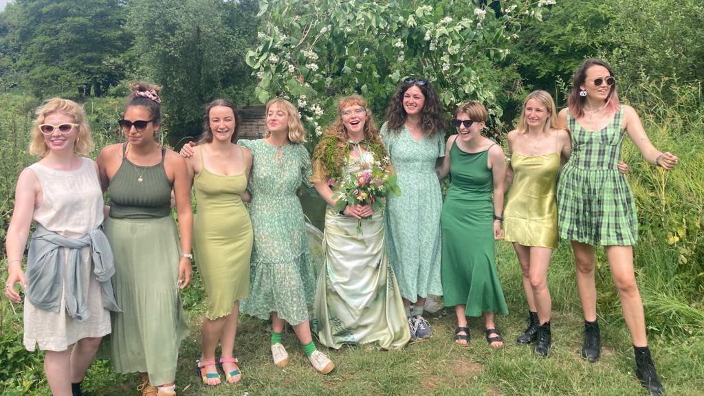 Megan Ruth-Trump stands in the centre of a group of eight wedding guests, all lined up in a row. She is wearing a wedding dress and holding a bouquet of flowers. The eight guests are all wearing dresses or tops and skirts, most of which are green