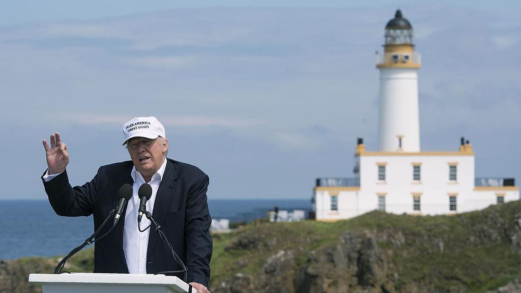 President Trump in white open-necked shirt and dark blue suit with white 'Make America Great Again' cap is holding up his right hand in a "zero" gesture. Behind and to his right is the lighthouse at Turnberry. The sky is bkue and we can see the sea.