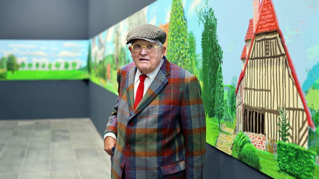 David Hockney, i veshur me syzet e tij karakteristike të rrumbullakëta me një kapelë të sheshtë, xhaketë me katrorë dhe kravatë të kuqe me këmishë të bardhë, qëndron përpara një vepre arti që shtrihet në një panel rreth dhomës ku ndodhet ai - në stilin tipik të Hockney-t, ajo përshkruan ndërtesa dhe fshatrat e Normandisë.
