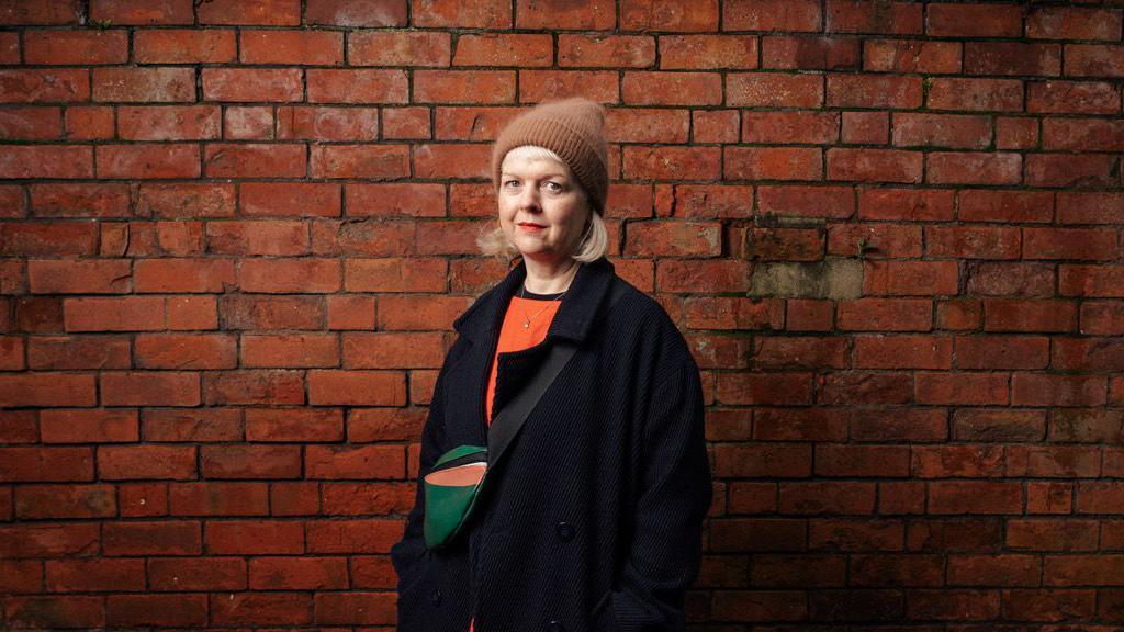 Amberlea is standing in front of a red brick wall. She is looking straight at the camera and is wearing a brown hat. She has short blonde hair and an orange jumper with a navy overcoat. 