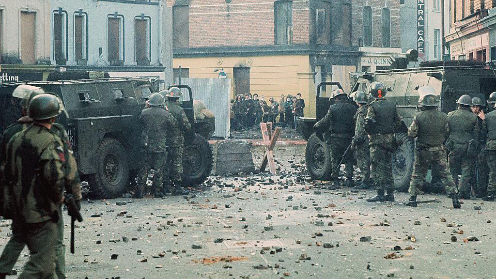 Two black army jeeps are parked at a barricade on a street in Londonderry. There are soldiers behind the jeeps taking shelter, they are armed and wearing camouflage clothing and hard hats. In the distance there is a group of young people. There is stones and other debris scattered across the street.