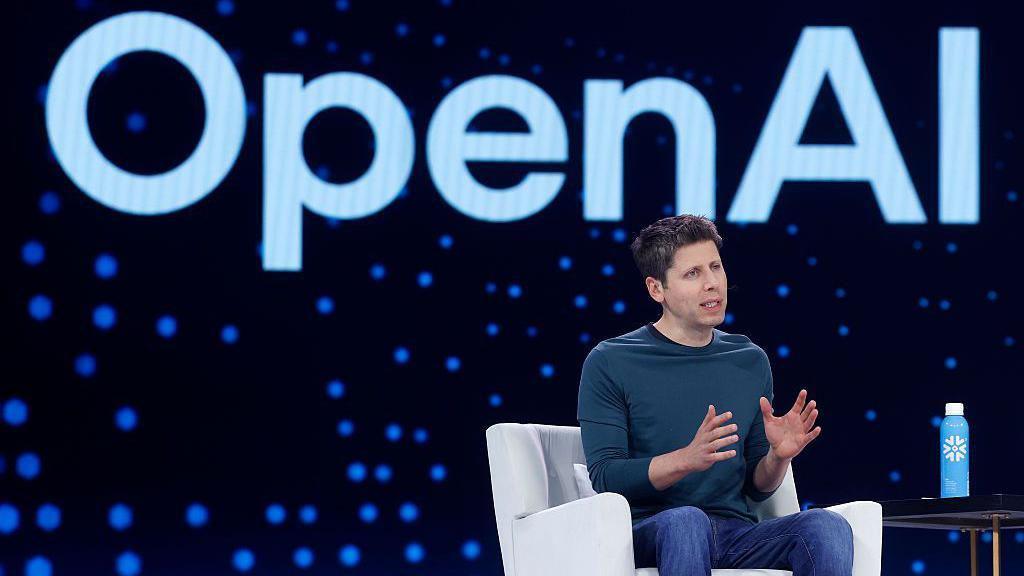 Open AI CEO Sam Altman speaks during Snowflake Summit 2025 at Moscone Center on June 02, 2025 in San Francisco, California. He is seen sitting on a white arm chair, wearing a dark long-sleeved dark teal shirt and blue denim jeans, gesturing with his hands as he speaks towards a crowd (not pictured). Behind him is a large electronic screen displaying the OpenAI logo.