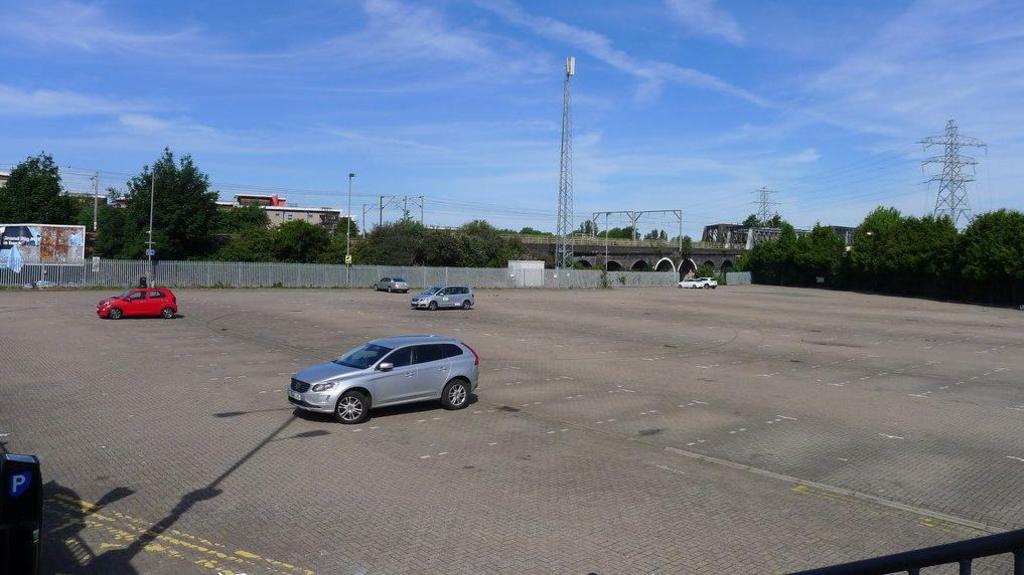 A mostly empty car park with four cars parked spread apart. Along the far edge is a metal gate and in the distance there are silver telephone masts and pylons in front of a blue sky.