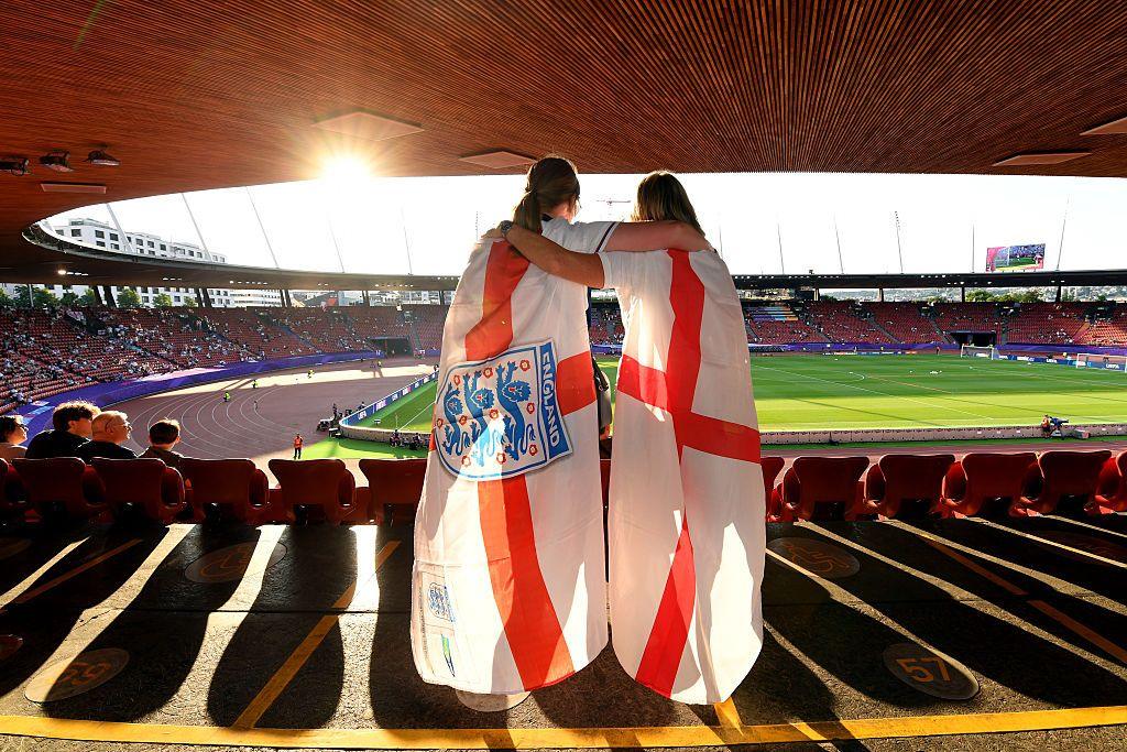 A back shot of two England fans wearing English flags over their shoulders and wrapping their arms around each other