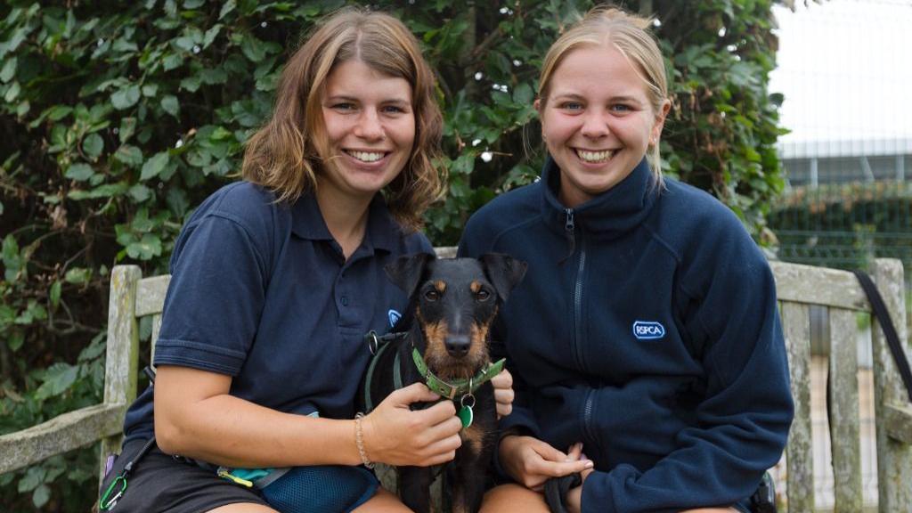 Two young women sit smiling on the bench, both with their hands resting on a small terrier-style dog that is resting between them. One of them has an RSPCA-branded top on and both are smiling at the camera