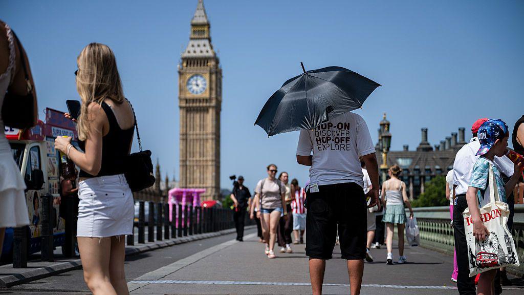 People in shorts and t shirts stand in the heat in London. Big Ben's tower can be seen in the background. 