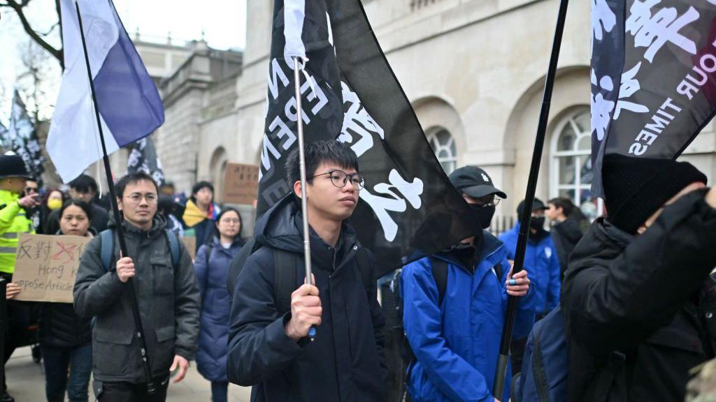 Tony Chung wearing a blue jacket holding a Hong Kong Independence flag surrounded by dozens of protesters holding pro-democracy flags and placards.