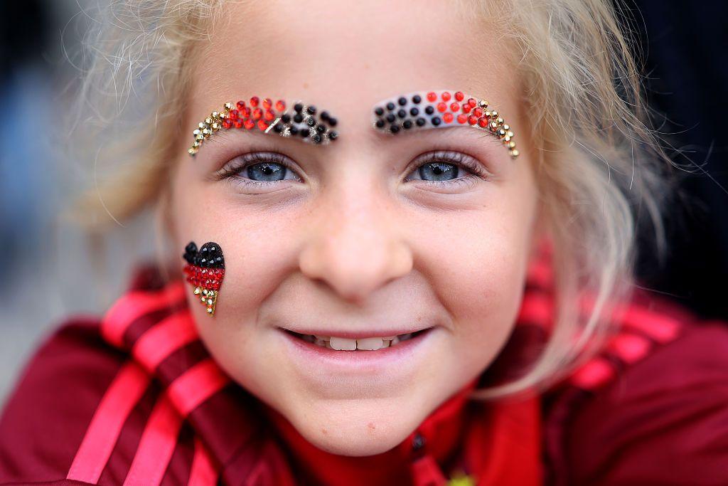 A young girl with blonde hair and blue eyes smiles to the camera in a close up shot. She is wearing sparkly eyebrows made out of beads in the colours of the German flag and a little heart on her right cheek