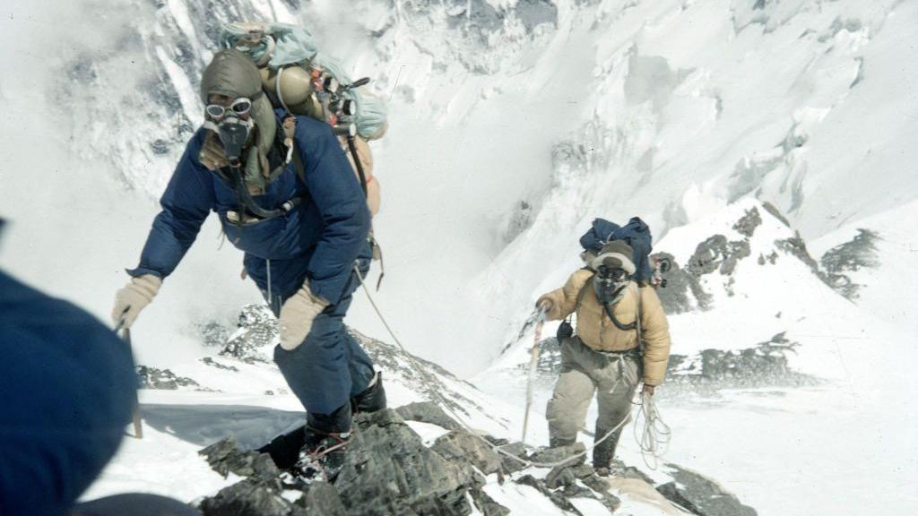 Edmund Hillary and Tenzing Norgay climbing. Hillary is dressed in blue padded trousers and top, with white mittens, and is wearing an oxygen mask and a pilot style leather helmet, behind him Norgay is in a yellow padded jacket with an oxygen mask and helmet. Both are wearing large rucksacks and the pair are roped together in the snow.