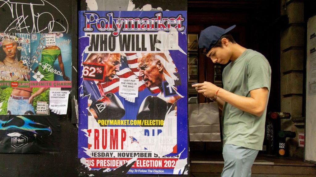 A young man looking at his phone walks past a ripped up advertisement for betting site Polymarket in the Brooklyn borough of New York, US, from July 2024 that asks, "Who will win? Trump v Biden"